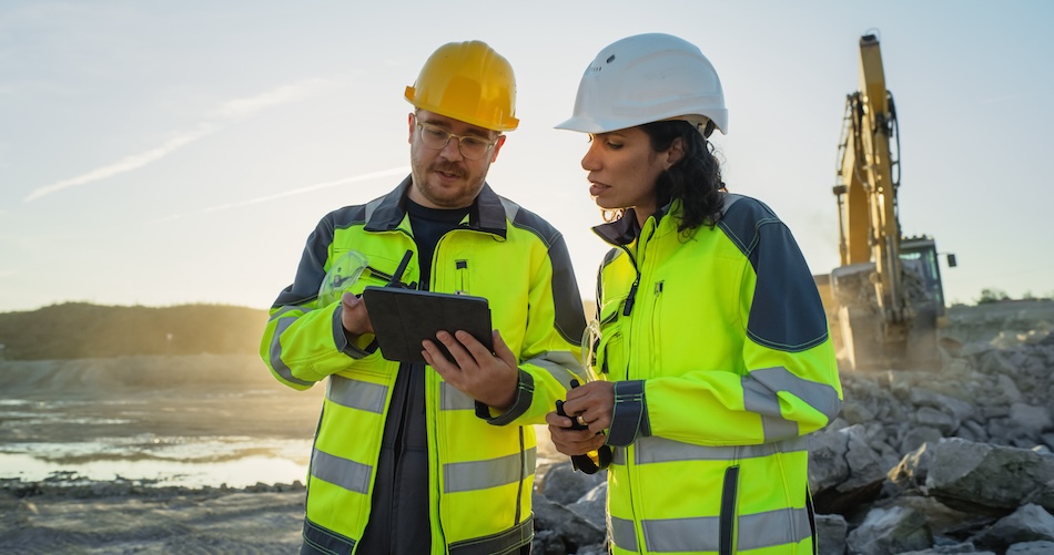 Caucasian Male Civil Engineer Talking To Hispanic Female Inspector And Using Tablet On Construction Site of New Apartment Building. Real Estate Developers Discussing Business, Excavators Working.