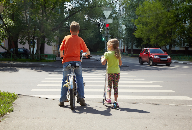Boy with bicycle and his younger sister with scooter stand before crossing at red traffic light